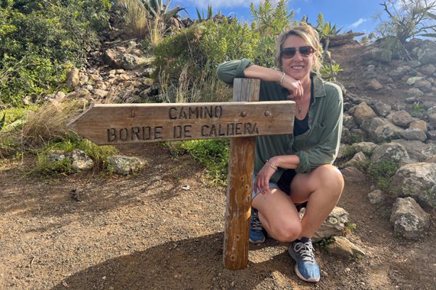 A blonde woman in sunglasses and shorts leaning on a wooden sign post